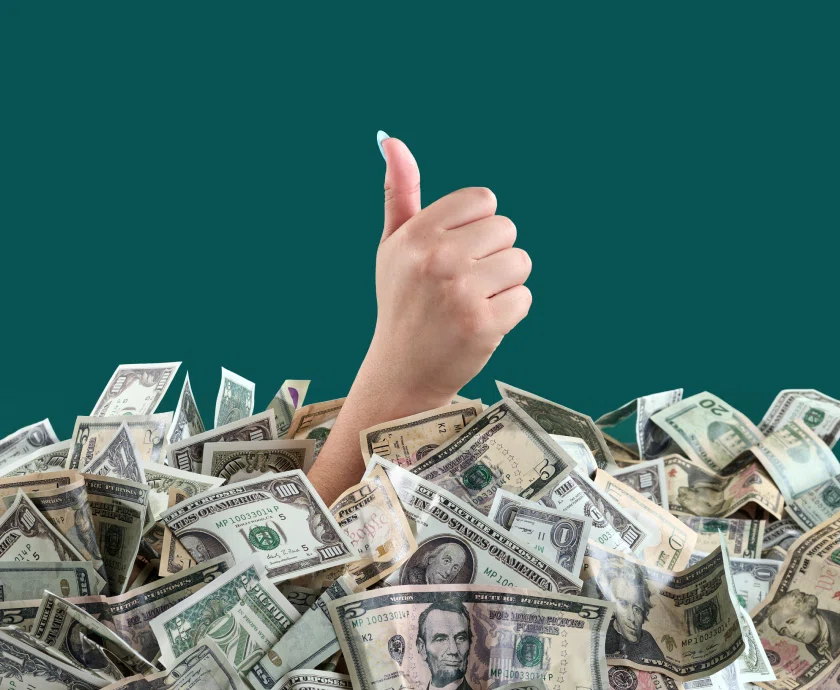 A hand giving a thumbs-up emerging from a large pile of various U.S. dollar bills, set against a solid green background.