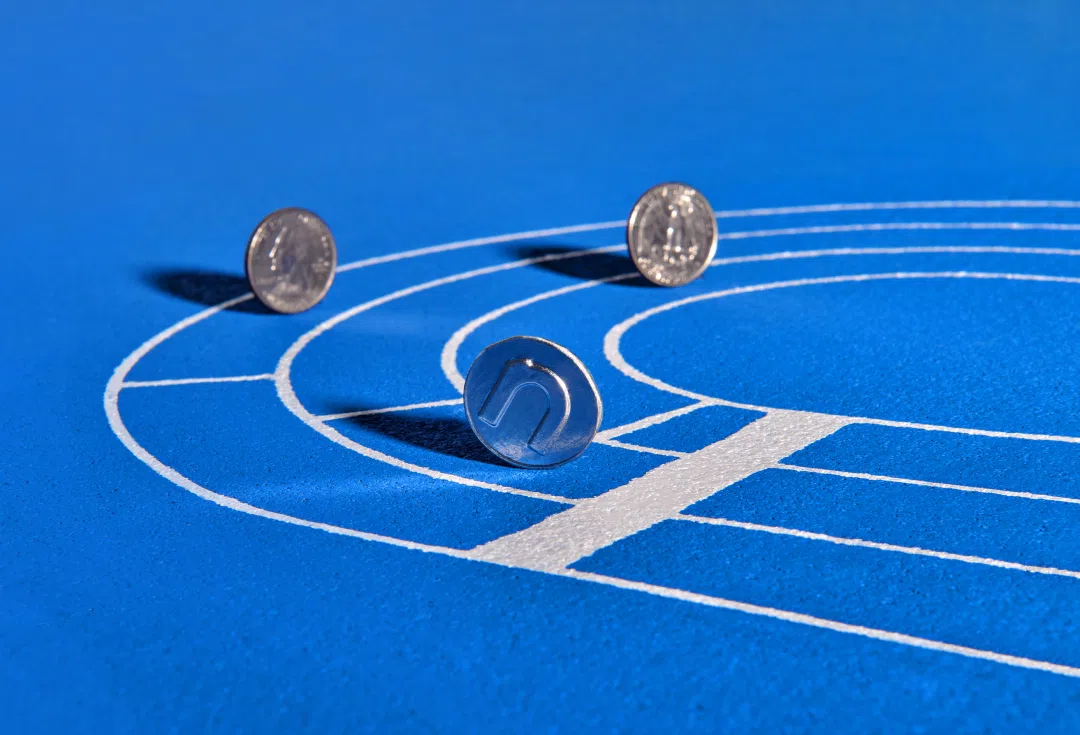 Three coins positioned on a blue running track with white lane markings. One coin is rolling, while the other two are standing on their edges.