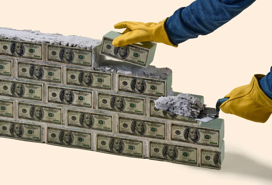 Concrete bricks made of U.S. dollar bills, forming a small wall, set against a solid peach background.